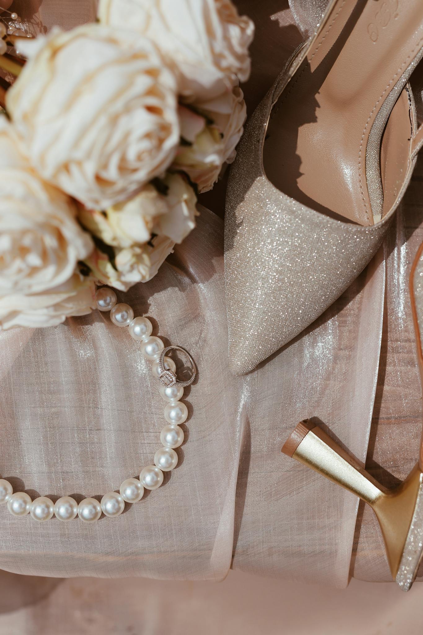Flat lay of elegant wedding shoes, pearl necklace, and roses on satin fabric.